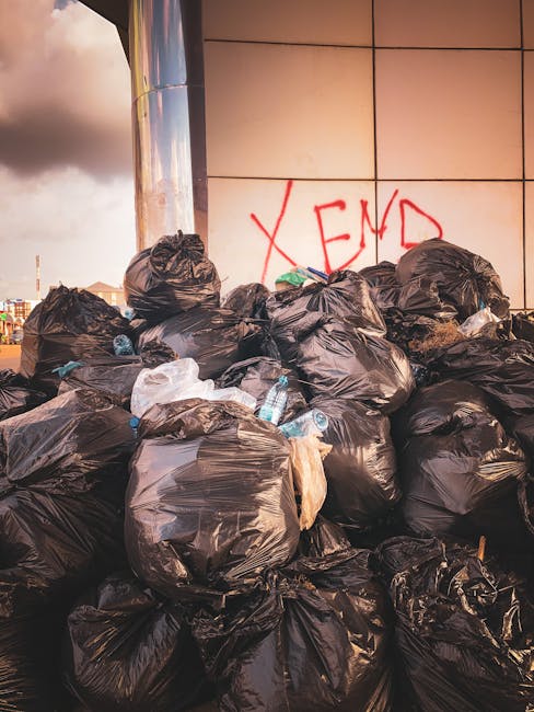 A large pile of black plastic rubbish bags filled with waste, stacked against a tiled wall with red graffiti spelling 'XEND'. Some of the bags are torn open, revealing plastic bottles and other household trash inside. The scene is outdoors, with a cloudy sky visible in the background. The image captures a waste collection point, highlighting the importance of proper disposal in relation to house removals and bulk waste management services provided by companies like Man with Van Kenley. The setting emphasizes the need for efficient rubbish removal during home relocation or when complying with local waste collection regulations, as discussed in the page about avoiding Croydon Council fines and bulky waste rules in Kenley.