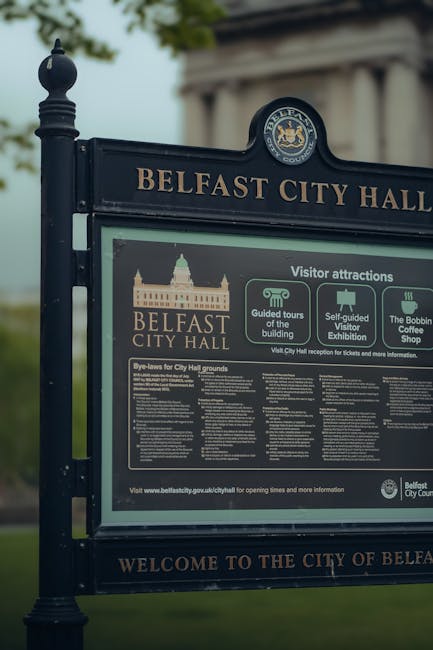 A close-up of a black metal signboard for Belfast City Hall, featuring the official city crest at the top. The sign displays information about visitor attractions, including guided tours of the building, self-guided visitor exhibitions, and The Bobbin Coffee Shop. Below, there is a section with bye-laws for City Hall grounds written in white text on a black background. The sign is situated outdoors, with a blurred background of green foliage and a classical stone building with columns partially visible. The sign is clean, well-maintained, and positioned on a paved surface, likely near an entrance or public area associated with Belfast City Hall. The overall scene is illuminated by natural daylight.