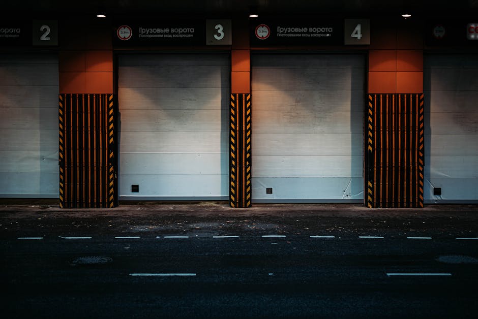 A large industrial warehouse loading bay with four closed roller shutter doors, numbered 2, 3, and 4, at night. The doors are flanked by black and yellow striped protective barriers. Above the doors, signage in a foreign language indicates loading zones or restricted access areas. The exterior wall is made of grey concrete panels, and the street in front has white dashed lines marking lanes. The area is illuminated by overhead lighting, casting shadows on the ground. This setting illustrates a typical logistics and freight handling environment, relevant to home relocation and furniture transport services provided by Man with Van Kenley, with an emphasis on the loading process in commercial or residential building access points.
