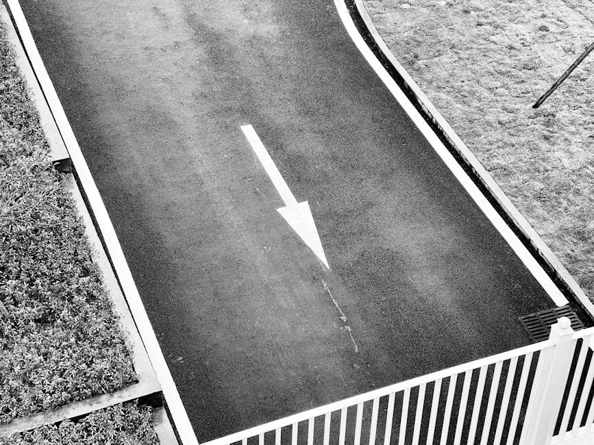 A black asphalt driveway with a large white directional arrow painted in the center, indicating the designated direction for vehicle movement. The driveway is bordered by a white picket fence on one side and a strip of soil with a pole or stake near the edge. Adjacent to the driveway, there is a small section of neatly trimmed greenery. The area appears well-maintained and is part of a residential property, facilitating the loading process for a home relocation. The scene is well-lit, capturing the smooth surface of the driveway and the clear markings used to manage traffic flow during furniture transport and packing and moving operations, with occasional reference to local house removals services such as those offered by Man with Van Kenley.