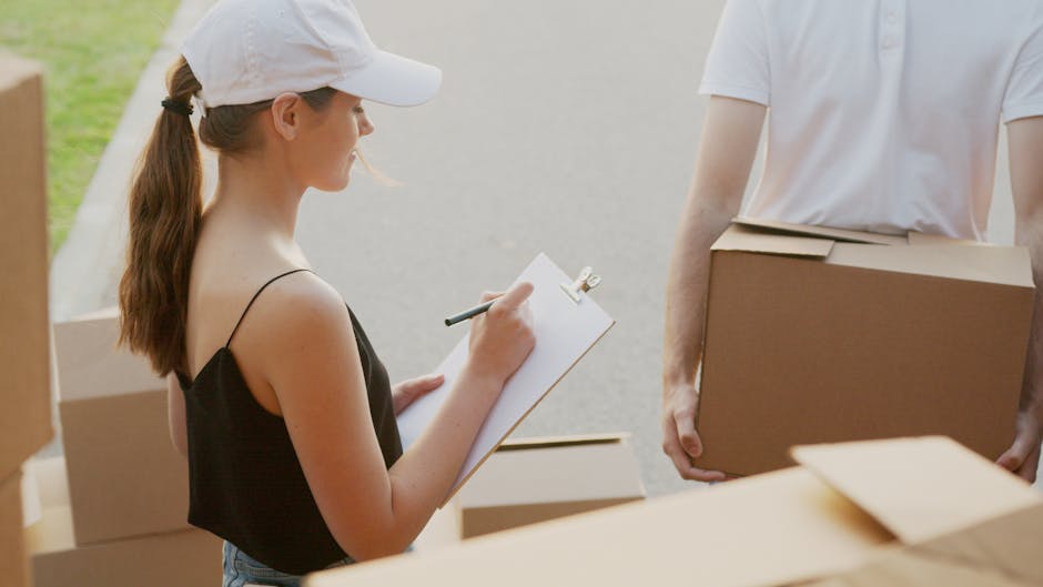 A close-up of a person's arm holding a black marker and writing on a large cardboard box labeled 'KITCHEN' with a small heart symbol. The box is part of a collection of similarly sized cardboard boxes