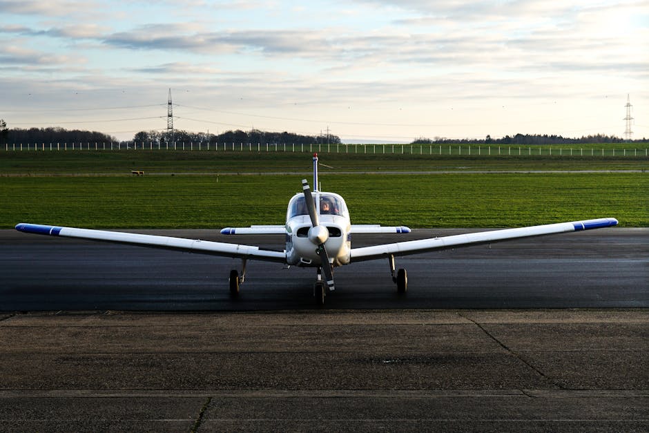 A small white aircraft parked on the tarmac at Kenley Aerodrome during daylight hours, with its nose pointed directly toward the camera. The aircraft features a sleek fuselage, a single propeller at the front, and a clear cockpit canopy revealing the interior. Its wings extend horizontally on either side, with blue accents on the wing tips. The aircraft's landing gear is visible, with the wheels resting on the asphalt surface. Behind the plane, there is a grassy field that stretches toward the horizon, bordered by a line of trees and a few utility poles with power lines. The sky above is partly cloudy with a soft, diffused light illuminating the scene. This image captures the quiet, functional environment of a small regional aerodrome, illustrating the type of location where Man with Van Kenley might organise logistics for vehicle or equipment transport related to house removals or relocation services at Kenley Aerodrome.