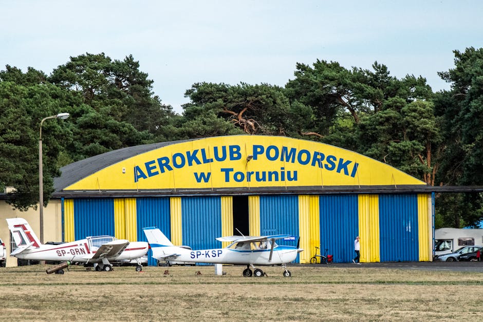 A large, curved hangar with a blue and yellow exterior, prominently displaying the sign 'AEROKLUB POMORSKI w Toruniu' on its façade. In front of the hangar, three small light aircraft are parked on a grassy area, with two featuring white fuselages accented by blue and red markings. The aircraft are positioned in a row, with the tail sections pointing to the left and the entry doors toward the hangar. The scene is set during daytime with diffuse natural lighting, and tall green trees border the background behind the hangar, creating a natural backdrop. To the right of the aircraft, a person is seen standing next to a bicycle, near the hangar entrance, which appears open. The environment suggests a general aviation facility or flying club, with an emphasis on light aircraft and hangar-based operations, consistent with home relocation or equipment transport services offered by Man with Van Kenley at this site.