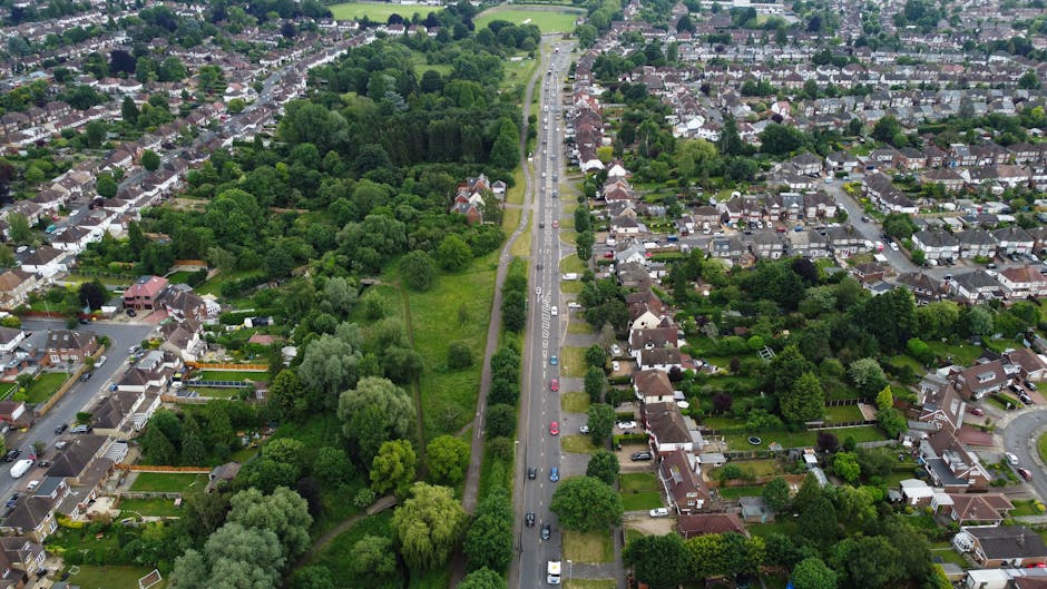Aerial view of a residential area during daytime showing a main road with cars parked along both sides, flanked by houses with gardens and a lush green park with mature trees and open grassy areas on the left. The houses are primarily detached and semi-detached, with visible driveways, backyards, and some garden furniture. The scene includes visible pavement, greenery, and the edges of a woodland area, illustrating a typical suburban neighbourhood suitable for house removals and furniture transport. The lighting indicates clear weather, with sunlight casting shadows on the trees and buildings. This image captures the environment and logistics involved in local home relocation, highlighting the importance of planning routes through residential streets and green spaces, as managed by companies like Man with Van Kenley.