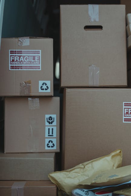 A close-up view of several cardboard boxes used for home relocation, stacked inside a property near a doorway. The boxes vary in size and feature printed handling labels, including a 'fragile' warning