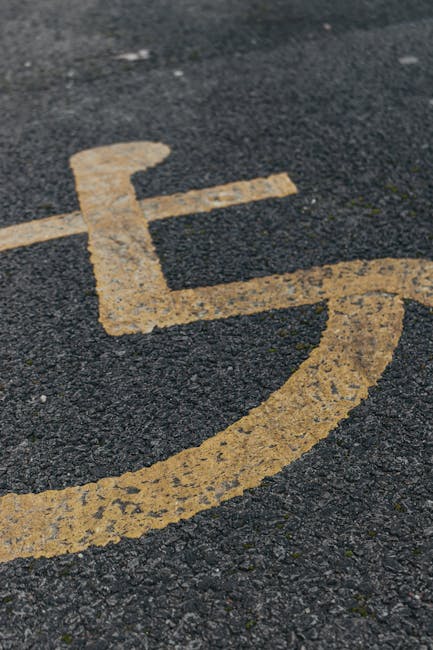 A close-up view of a yellow wheelchair symbol painted on a dark asphalt surface, indicating a designated accessible parking space. The paint appears weathered and slightly worn, with some areas showing chipping and minor deterioration. The image captures only the pavement and the wheelchair icon, with no other objects or vehicles visible in the frame. This visual is relevant for illustrating accessible parking provisions often associated with residential or commercial building entrances, which are important considerations during home relocations or professional removals. Man with Van Kenley occasionally incorporates such context in their content about moving near Kenley Station, highlighting parking access and logistical planning for furniture transport and packing during house moves.
