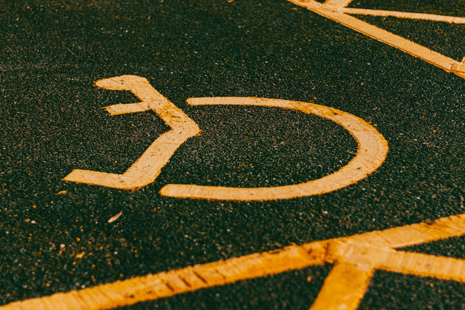 Close-up image of a yellow disabled parking bay marking painted on an asphalt surface, with a focus on the wheelchair symbol. The textured asphalt shows small stones and rough patches, with some faint yellow lines marking adjacent parking spaces. The marking appears slightly worn but still clearly visible. This parking area is near a property or street, likely used by visitors or residents. The image demonstrates the type of accessible parking facilities commonly found near homes, businesses, or public buildings, relevant to home relocation and moving services provided by Man with Van Kenley for efficient access and parking considerations during furniture transport and packing tasks.