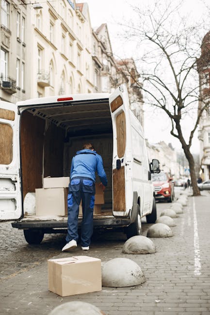A man dressed in blue work clothes is loading or unloading a large white moving van parked on a city street. The van's rear doors are open, revealing empty wooden crates and packed cardboard boxes inside. The man is bending forward, handling items on the floor of the van, possibly during a home relocation or furniture transport process. In front of the van, a cardboard box is placed on the pavement, which is lined with round concrete bollards separating the pedestrian area from the roadway. The street features traditional European-style buildings with multiple windows and ornate facades, with a leafless tree in the background indicating a winter or early spring season. Several parked cars are visible along the street, and the overall scene depicts a professional removal or moving service. This photo is associated with Man with Van Kenley, offering quick and efficient removals that include packing, loading, and transportation services.