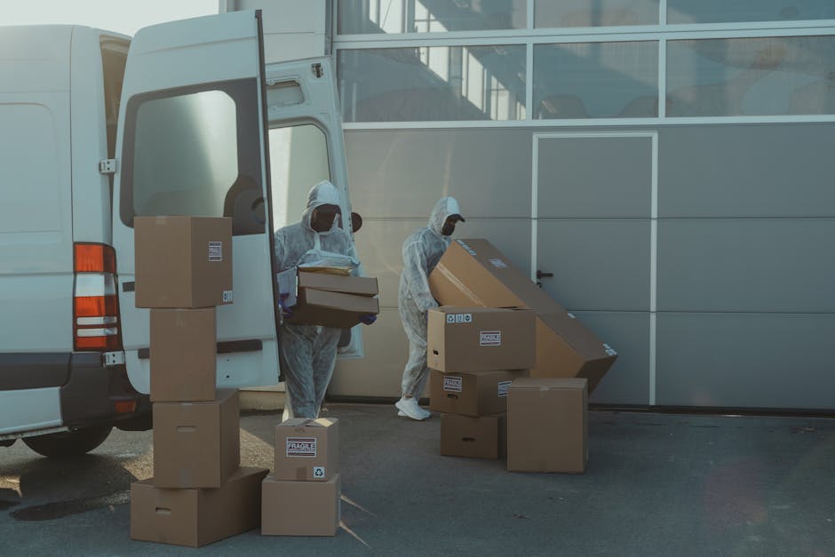 Two workers dressed in full protective personal equipment, including white suits and face masks, are engaged in a home relocation process outside a modern building with large glass windows. They are unloading and carrying brown cardboard boxes of various sizes from a white panel van, which is parked with its side door open. One worker is lifting a medium-sized box, while the other is positioning a large, flat box to be moved. Several packed boxes are stacked nearby, some labeled with fragile stickers. The scene includes a wheeled trolley and moving blankets on the ground, indicating a professional furniture transport and packing and moving operation by Man with Van Kenley. The background features a sleek, grey metallic garage door, and the overall environment is well-lit, suggesting daytime conditions. The image captures the logistics involved in a house removal, emphasizing careful handling of packed items and organized loading procedures.