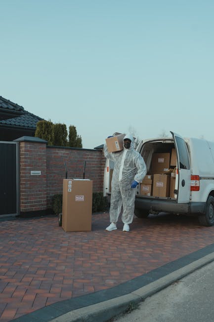 A man dressed in blue work clothes is loading or unloading a large white moving van parked on a city street. The van's rear doors are open, revealing empty wooden crates and packed cardboard boxes inside. The man is bending forward, handling items on the floor of the van, possibly during a home relocation or furniture transport process. In front of the van, a cardboard box is placed on the pavement, which is lined with round concrete bollards separating the pedestrian area from the roadway. The street features traditional European-style buildings with multiple windows and ornate facades, with a leafless tree in the background indicating a winter or early spring season. Several parked cars are visible along the street, and the overall scene depicts a professional removal or moving service. This photo is associated with Man with Van Kenley, offering quick and efficient removals that include packing, loading, and transportation services.