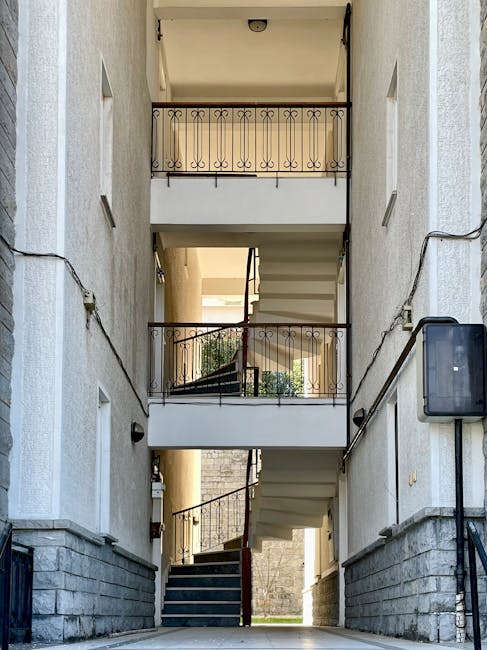 The image depicts a multi-storey residential building with a narrow, enclosed staircase and balconies on each level, viewed from the ground floor. The staircase is partially visible, winding upwards behind a white wall, with a metal handrail along the corridor. Balconies with decorative black wrought iron railings are situated on each floor above the stairwell. The ground floor area shows a paved surface, with a doorway leading into the building on the right, and a black electrical box mounted on the exterior wall. The interior walls are light-colored, and there are small rectangular windows in the walls adjacent to the staircase to allow natural light inside. This setting appears suitable for house removals or moving services, such as those provided by Man with Van Kenley, especially for transporting furniture or boxes through narrow entrances during a relocation process.