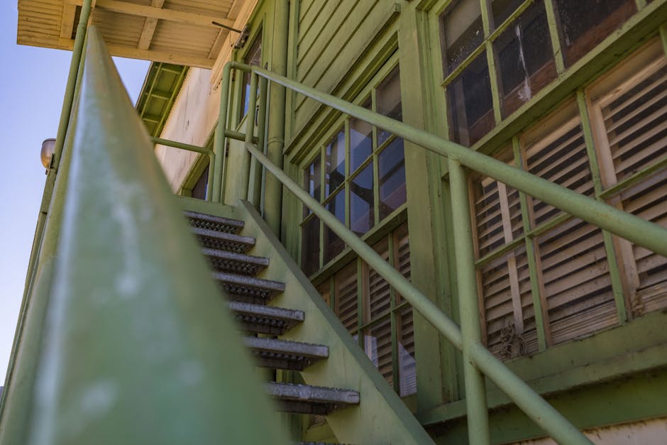 A view from the ground of a green-painted exterior staircase with metal treads leading up to a building with large windows, some with wooden blinds. The staircase has a matching green handrail on both sides, supported by vertical posts. The building's façade features wooden panels, some of which appear weathered or partially disassembled. Inside, visible through the window, there are cardboard boxes, packing materials, and possibly furniture components, indicating a home relocation process. The scene is illuminated by natural daylight, with clear skies overhead, and the area around the building appears to be part of a move managed by Man with Van Kenley, specialists in removals and furniture transport for challenging staircases and narrow entrances, ensuring smooth and secure loading for house moves.