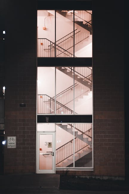 The image depicts a multi-storey residential building with a narrow, enclosed staircase and balconies on each level, viewed from the ground floor. The staircase is partially visible, winding upwards behind a white wall, with a metal handrail along the corridor. Balconies with decorative black wrought iron railings are situated on each floor above the stairwell. The ground floor area shows a paved surface, with a doorway leading into the building on the right, and a black electrical box mounted on the exterior wall. The interior walls are light-colored, and there are small rectangular windows in the walls adjacent to the staircase to allow natural light inside. This setting appears suitable for house removals or moving services, such as those provided by Man with Van Kenley, especially for transporting furniture or boxes through narrow entrances during a relocation process.